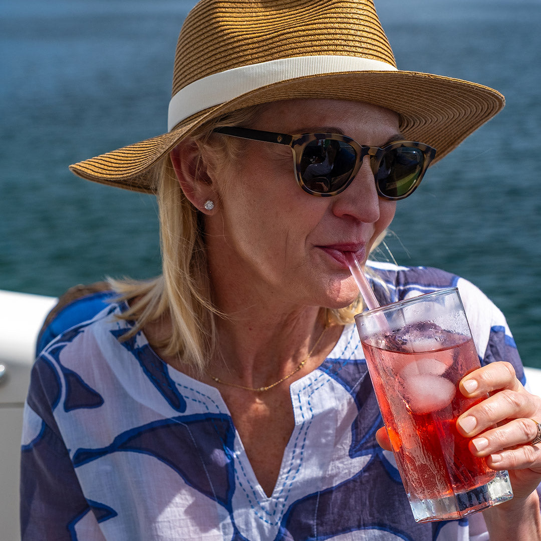 Woman wearing a straw hat and sunglasses, drinking from a glass of red tea with ice, against a blue water background.