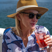 Woman wearing a straw hat and sunglasses, drinking from a glass of red tea with ice, against a blue water background.
