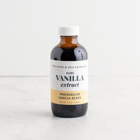 A bottle of Pure Vanilla Extract (Madagascar Vanilla Beans) from The Spice & Tea Exchange, featuring an amber glass bottle, white cap, and a white and beige label, set on a light marble surface against a white background.