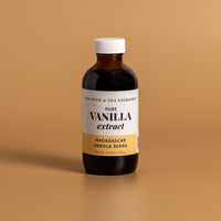 A bottle of Madagascar Vanilla Extract from The Spice & Tea Exchange, featuring a white label with a gold bottom section and a white cap, set against a solid tan background.