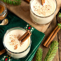 Two clear glasses filled with a creamy white beverage, garnished with cinnamon sticks and a sprinkle of brown spice, arranged on a rustic wooden table with pine branches, a green knit cloth, cinnamon sticks, and a small jar of spice.