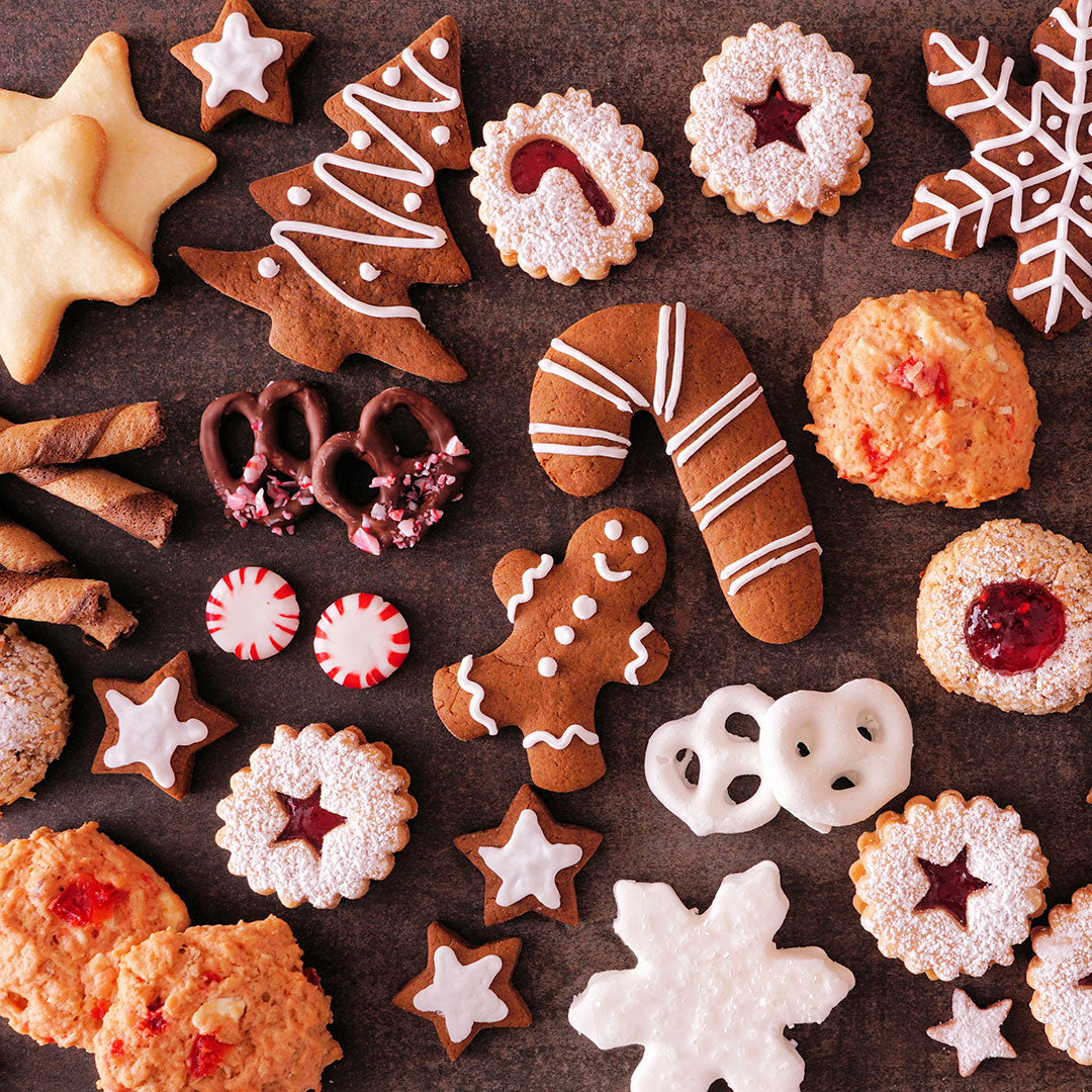 An overhead flat-lay of assorted holiday cookies and treats on a dark surface, including gingerbread men, Christmas trees, snowflakes, pretzels, candy canes, star-shaped cookies, and cookies with red fruit filling.