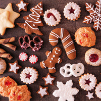 An overhead flat-lay of assorted holiday cookies and treats on a dark surface, including gingerbread men, Christmas trees, snowflakes, pretzels, candy canes, star-shaped cookies, and cookies with red fruit filling.