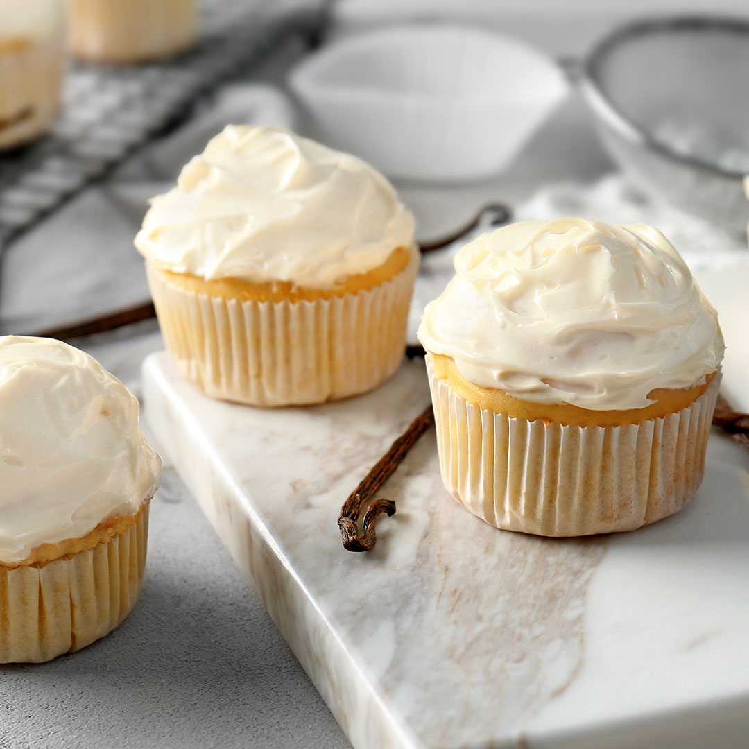 Three vanilla cupcakes with thick white frosting in paper liners, arranged on a white marble serving board with a vanilla bean, in a softly lit kitchen setting.