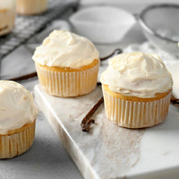 Three vanilla cupcakes with thick white frosting in paper liners, arranged on a white marble serving board with a vanilla bean, in a softly lit kitchen setting.