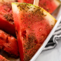 Close-up of watermelon slices on a stick, topped with a brown spice rub, arranged in a white dish with a striped cloth in the background.