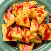 Close-up of triangular slices of pale yellow fruit, likely pineapple, tossed with red sauce and a brown spice blend, served on a green plate.