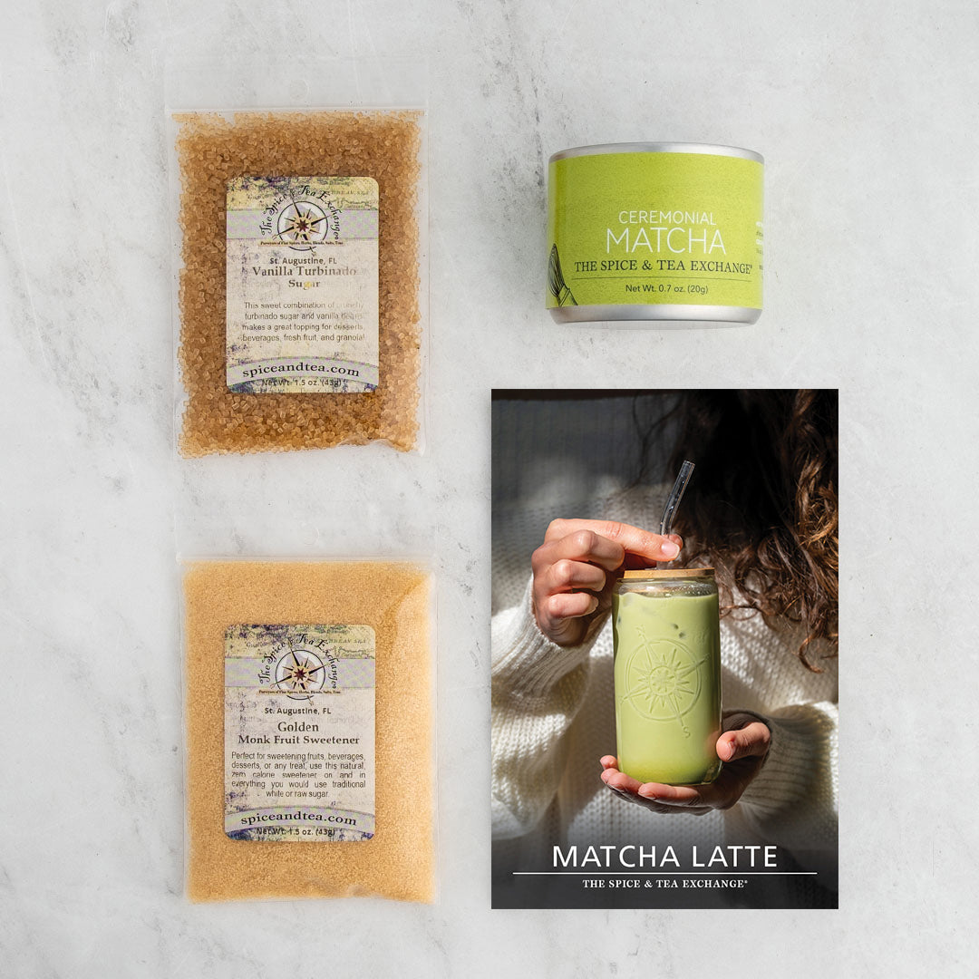 A flat lay of the Matcha Latte (Kit), featuring vanilla turbinado sugar, ceremonial matcha tin, golden matcha fruit sweetener, and a brochure with a hand holding a green matcha latte on a light-toned surface.