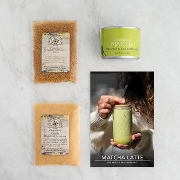 A flat lay of the Matcha Latte (Kit), featuring vanilla turbinado sugar, ceremonial matcha tin, golden matcha fruit sweetener, and a brochure with a hand holding a green matcha latte on a light-toned surface.