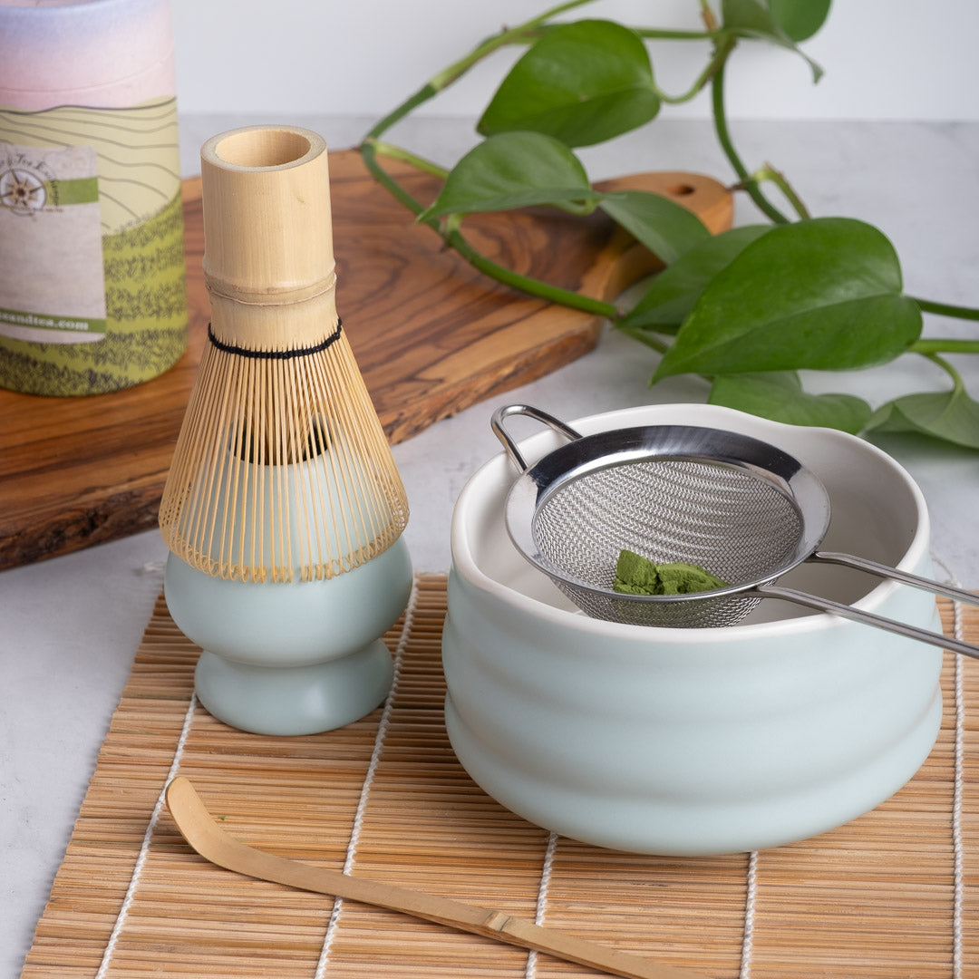 Tea-making setup with a Matcha Bowl (ceramic), Matcha Whisk (bamboo), Whisk Holder (ceramic), Matcha Sieve (stainless steel), Matcha Scoop (bamboo) on a wooden mat.