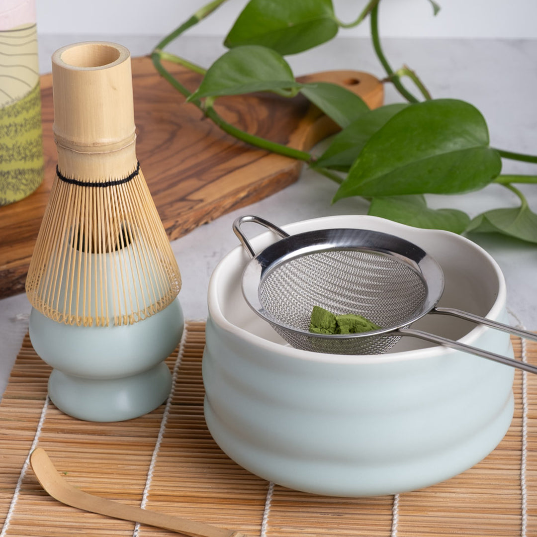 Tea-making setup with a bamboo whisk, ceramic bowl, and metal sieve on a wooden mat.