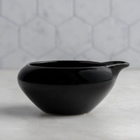 Black ceramic bowl with a handle on a light gray surface with a geometric patterned wall in the background.