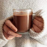 A person wearing a chunky cream knit sweater holds a clear glass mug filled with a warm brown beverage, shown from the chest up against a soft textured knit background.