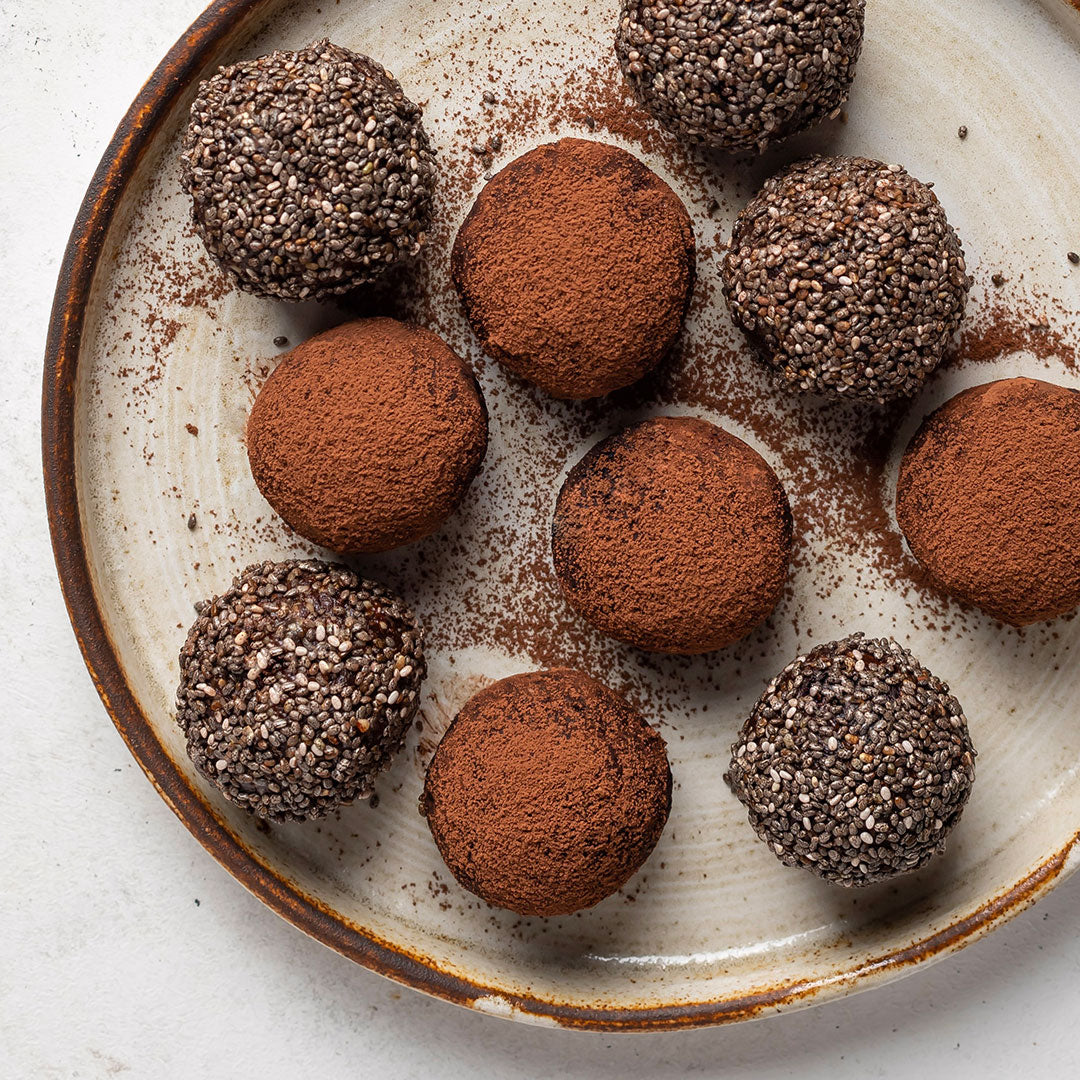 A top-down view of eight chocolate truffles, some coated in cocoa powder and others rolled in chia seeds, arranged on a rustic ceramic plate with cocoa powder scattered around them.