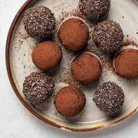 A top-down view of eight chocolate truffles, some coated in cocoa powder and others rolled in chia seeds, arranged on a rustic ceramic plate with cocoa powder scattered around them.