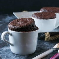 A white ceramic mug filled with a baked chocolate cake, set on a dark tabletop with two more mug cakes blurred in the background.