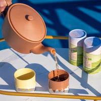 Person pouring tea from mocha mousse teapot into a matching cup on a blue and white striped surface.