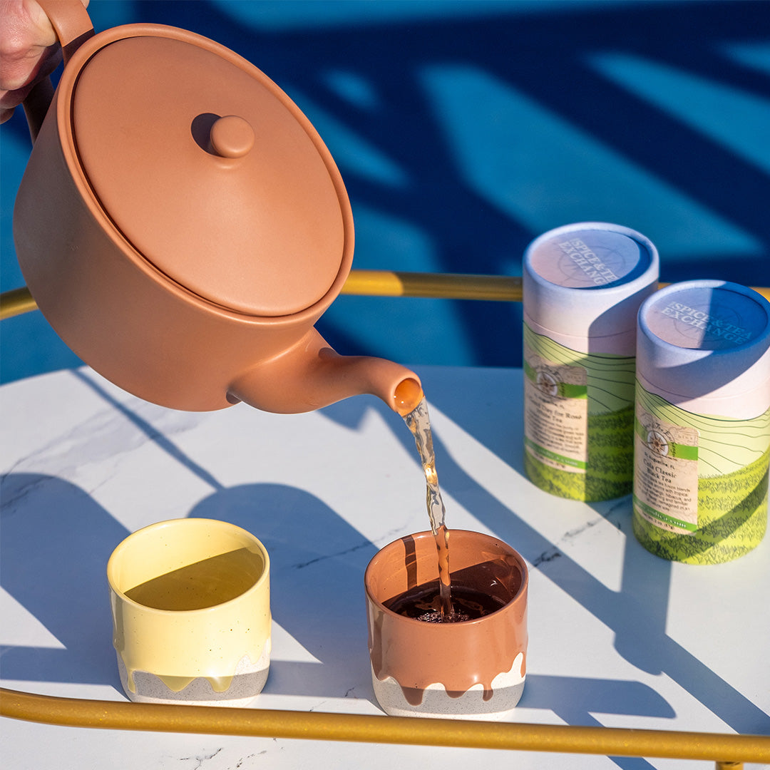 Person pouring tea from mocha mousse teapot into a matching cup on a blue and white striped surface.