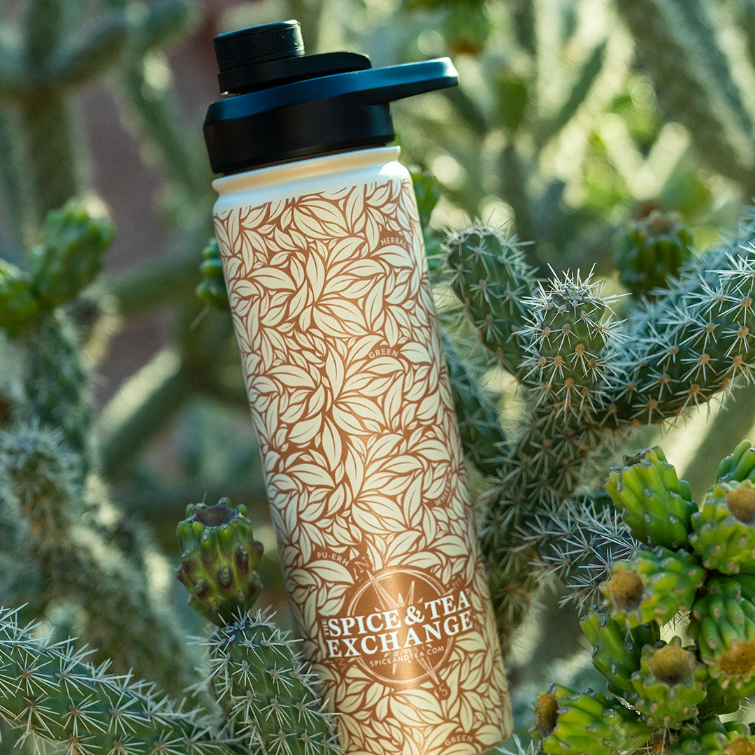 A reusable water bottle with a black flip-top lid and a beige body covered in a brown leaf pattern, standing upright among green cacti. The bottle is branded 'Spice & Tea Exchange'.
