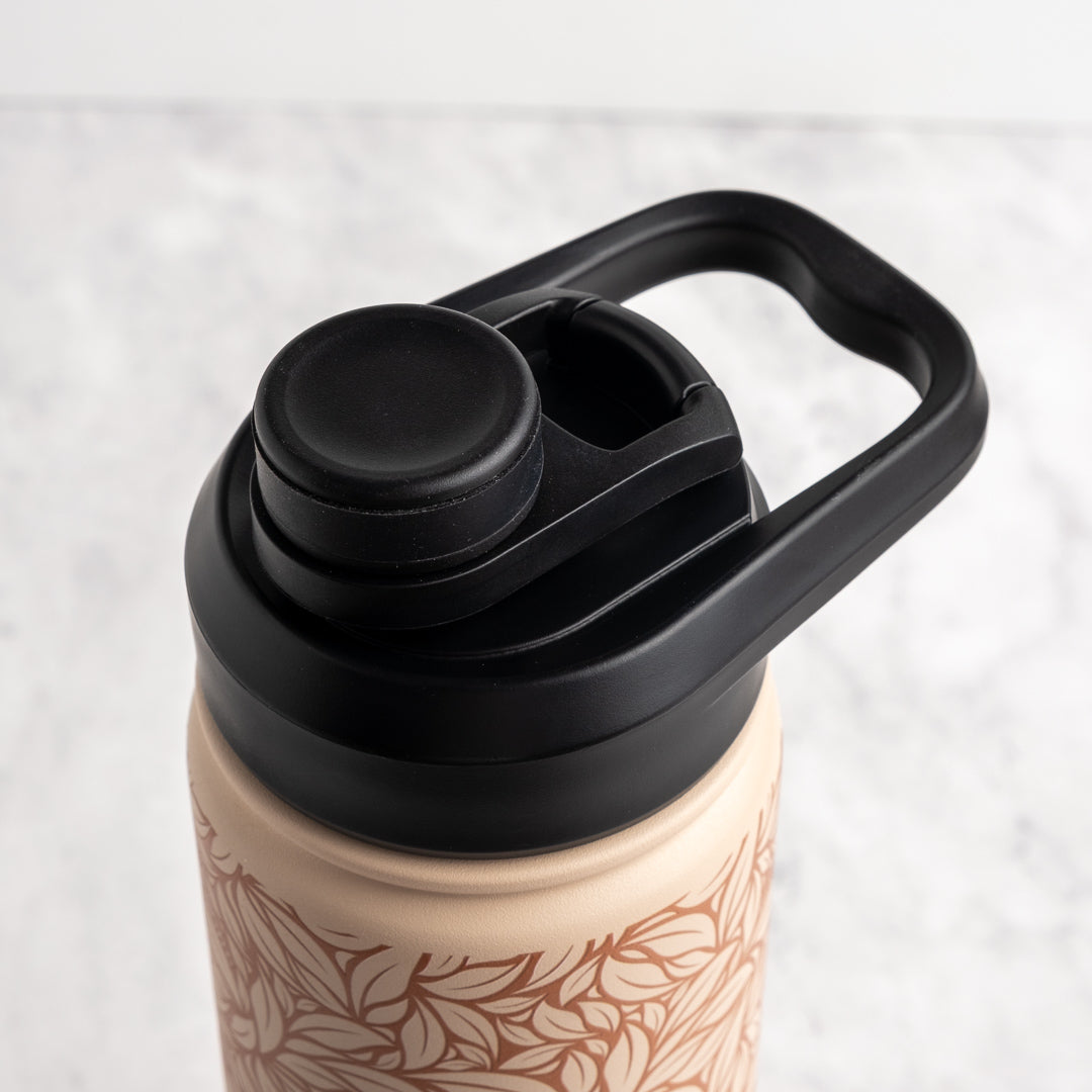 Close-up of a reusable water bottle with a black flip-straw lid and carrying loop, shown on a light marble-like surface. The bottle body is a warm beige with a brown leaf pattern.
