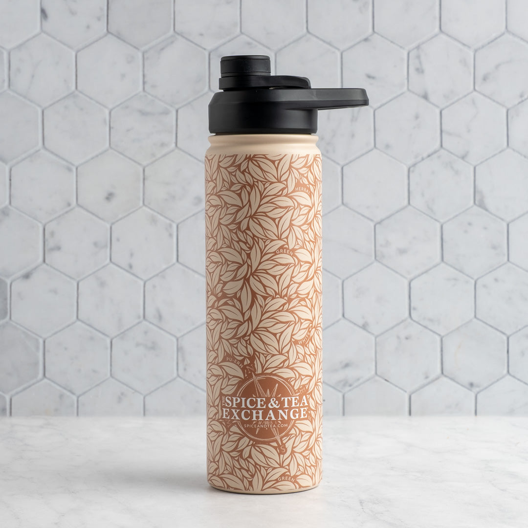 A reusable water bottle with a black flip-top lid, featuring a warm beige botanical leaf pattern and the 'Spice & Tea Exchange' logo, set against a light gray hexagonal tile background.