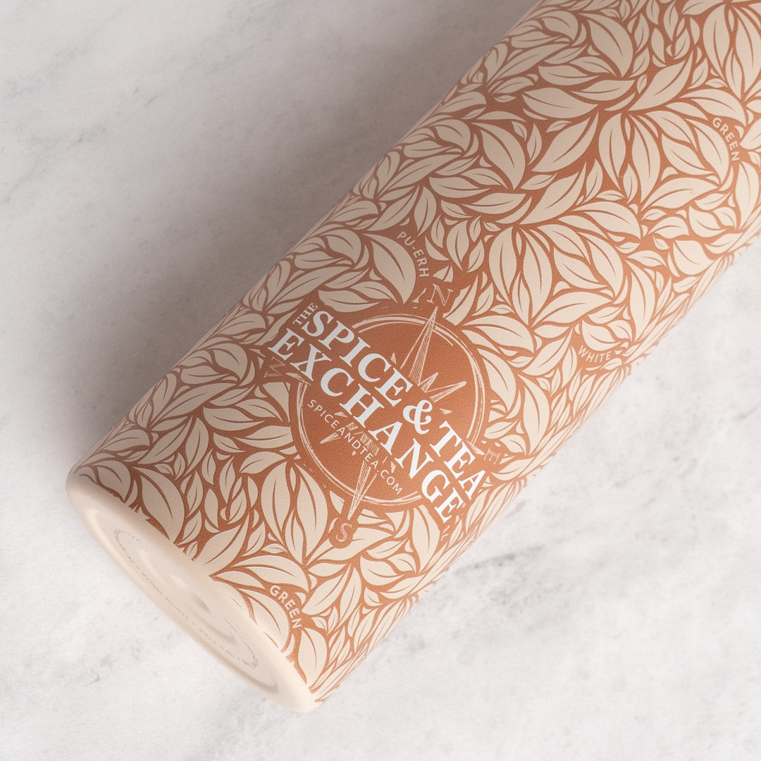 A cylindrical tumbler with a warm terracotta and cream botanical leaf pattern, featuring the 'Spice & Tea Exchange' logo in the center, resting on a light gray marble surface.