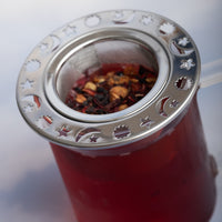 A glass mug of red herbal tea brewed with the Moon and Stars Infuser, featuring celestial cut-outs, holding aromatic loose-leaf tea and fruit pieces.