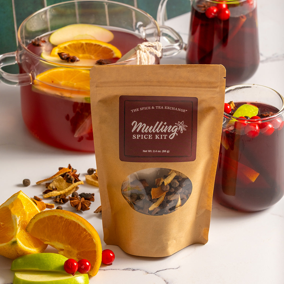 A kraft paper pouch labeled 'The Spice & Tea Exchange Mulling Spice Kit' stands on a white marble surface, surrounded by mulling spices, sliced orange, lime, and red berries, with mulling tea in glass pitchers in the background.