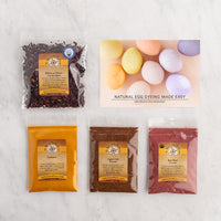 Four packaged natural dye ingredients and a printed recipe card are arranged on a light marble surface with the text 'Natural Egg Dyeing Made Easy'.