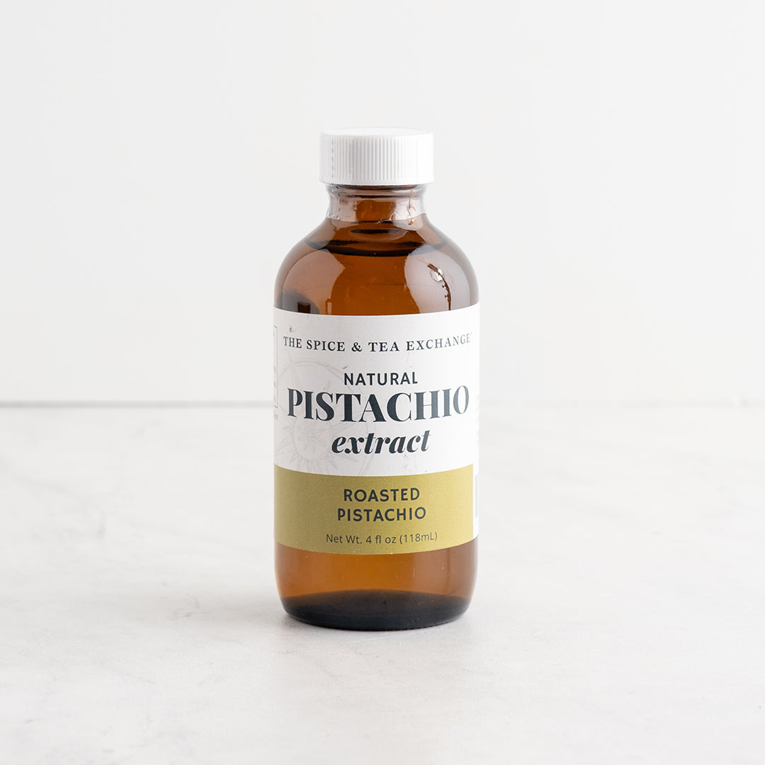 A 4 fl oz amber glass bottle of The Spice & Tea Exchange Natural Pistachio Extract, Roasted Pistachio flavor, with a white cap and a white and gold label, set on a light marble surface against a white background.