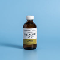 A 4 fl oz amber glass bottle of Natural Pistachio Extract by The Spice & Tea Exchange, featuring a white cap and a white label with a gold accent band, set against a solid light blue background.