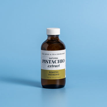 A 4 fl oz amber glass bottle of Natural Pistachio Extract by The Spice & Tea Exchange, featuring a white cap and a white label with a gold accent band, set against a solid light blue background.