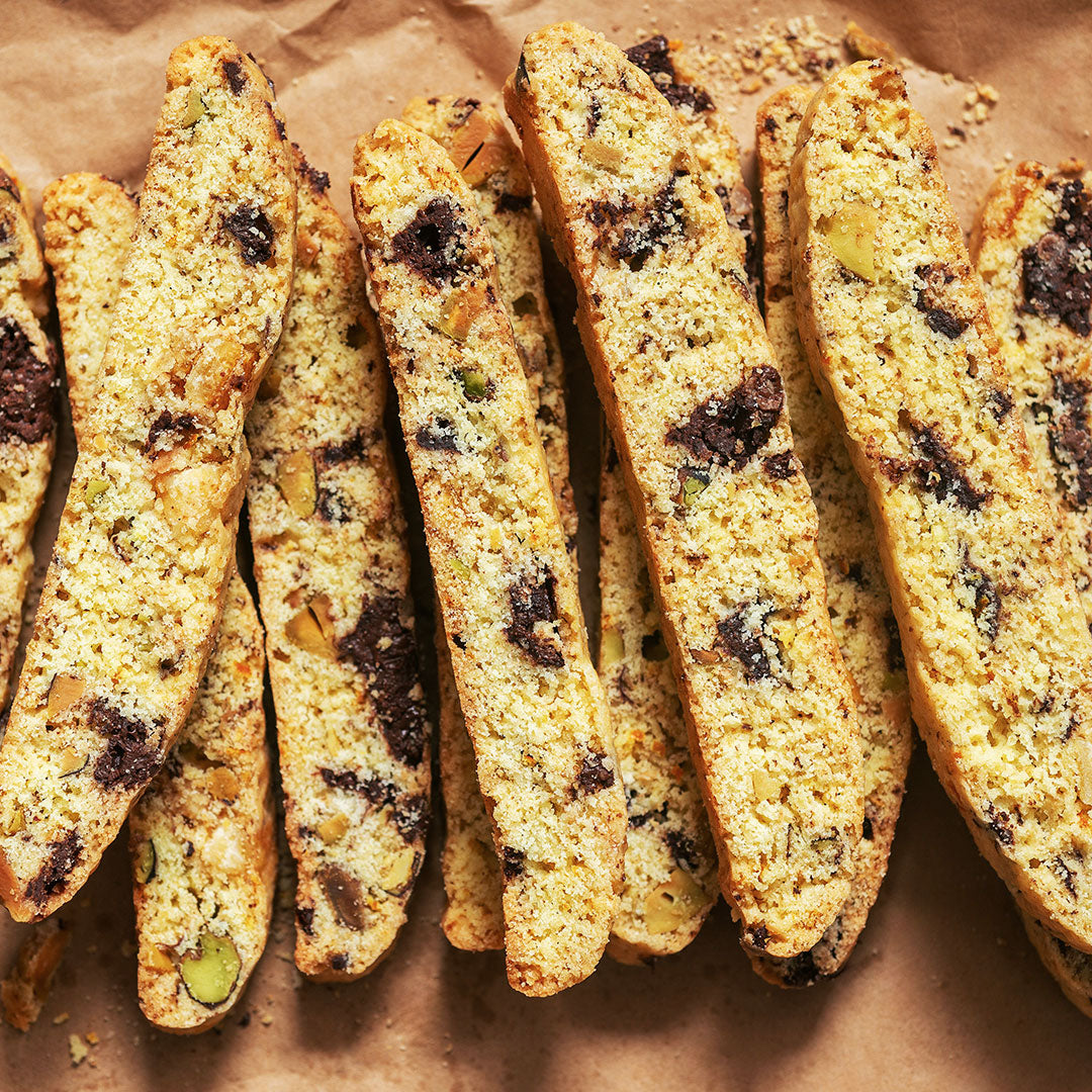 Close-up of several biscotti cookies arranged on brown parchment paper. The biscotti are golden tan with dark chocolate chips, nuts, and small green pieces visible inside.