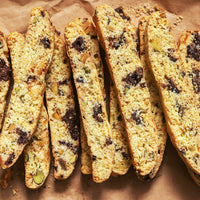 Close-up of several biscotti cookies arranged on brown parchment paper. The biscotti are golden tan with dark chocolate chips, nuts, and small green pieces visible inside.