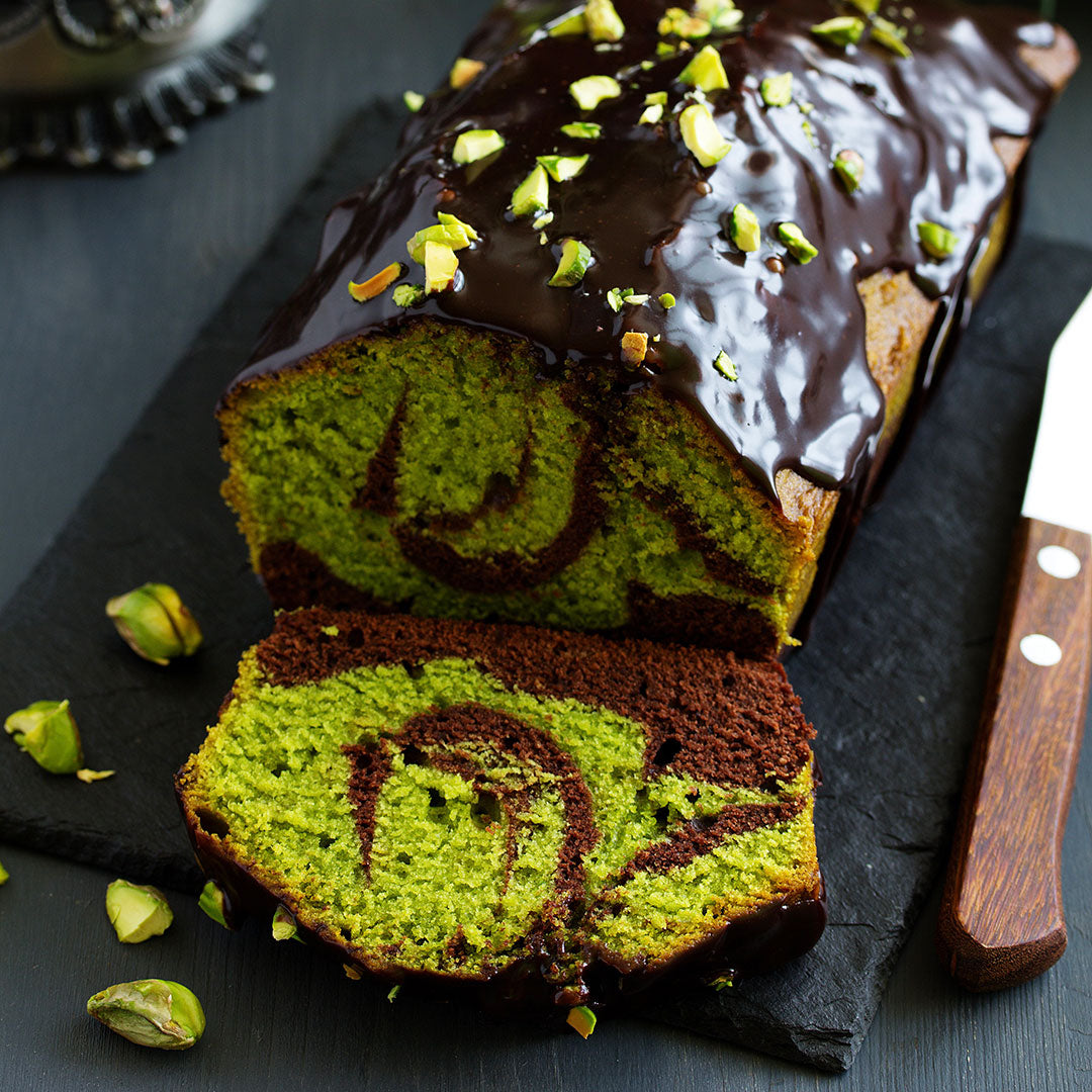 A sliced loaf of green and brown marble cake with glossy dark chocolate frosting and chopped pistachio pieces, presented on a dark slate board with a knife.