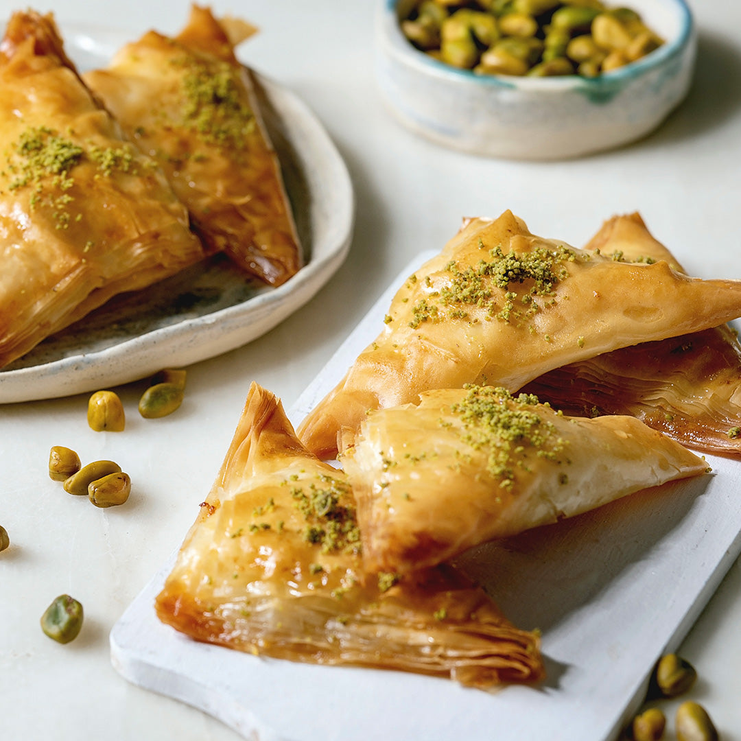Golden-brown flaky pastry triangles topped with chopped green pistachio, arranged on white serving boards with a small bowl of whole pistachios in the background.