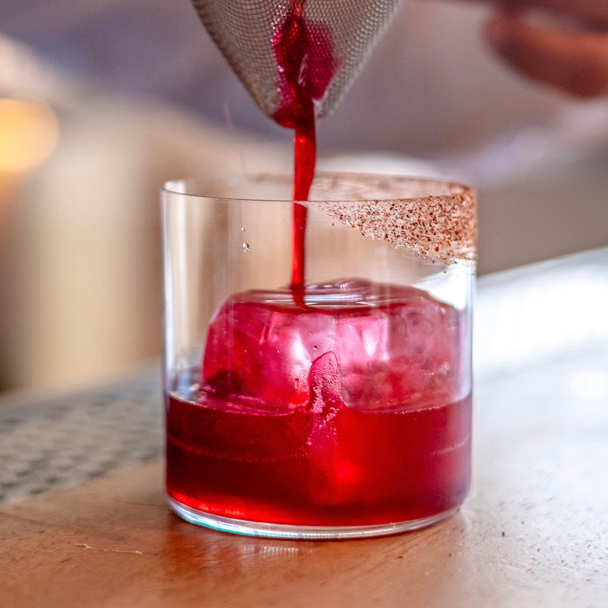 A close-up of a vibrant red cocktail being poured from a metal strainer into a clear glass tumbler with a sugar-coated rim, filled with ice on a wooden bar surface.