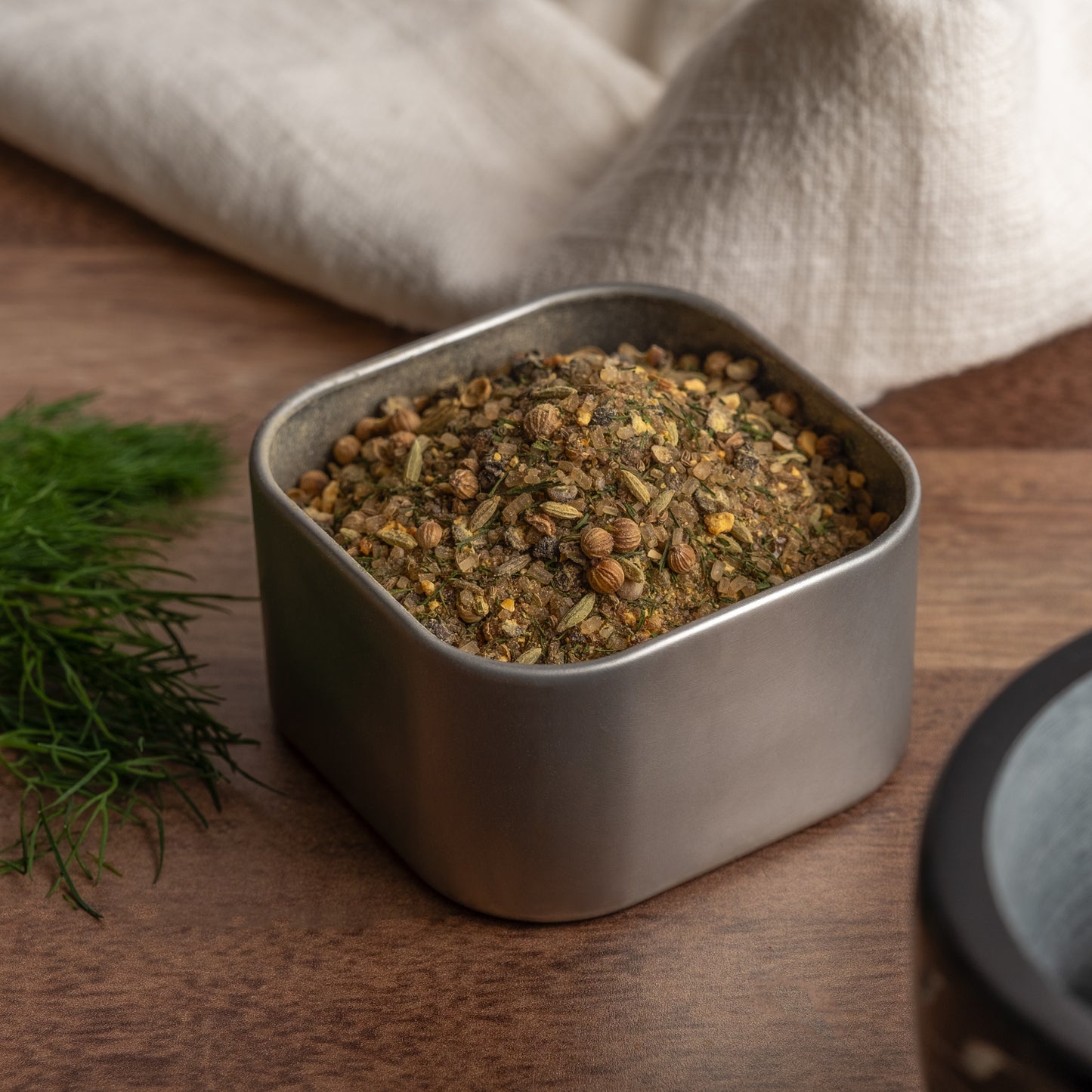 A square metal container of Northwest Salmon Rub, filled with a blend of dried herbs and gourmet fish seasonings, rests on a wooden surface. Fresh dill and a folded white cloth are in the background.