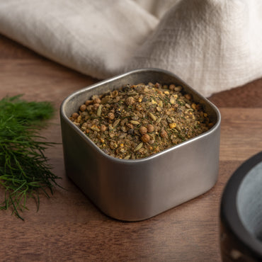 A square metal container of Northwest Salmon Rub, filled with a blend of dried herbs and gourmet fish seasonings, rests on a wooden surface. Fresh dill and a folded white cloth are in the background.