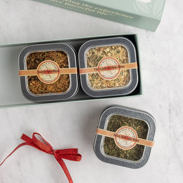 A gift set of three square metal spice tins displayed in and beside an open mint green box on a light marble surface. The tins have dark gray lids, clear windows showing brown and green spices inside, and tan paper bands with round labels.