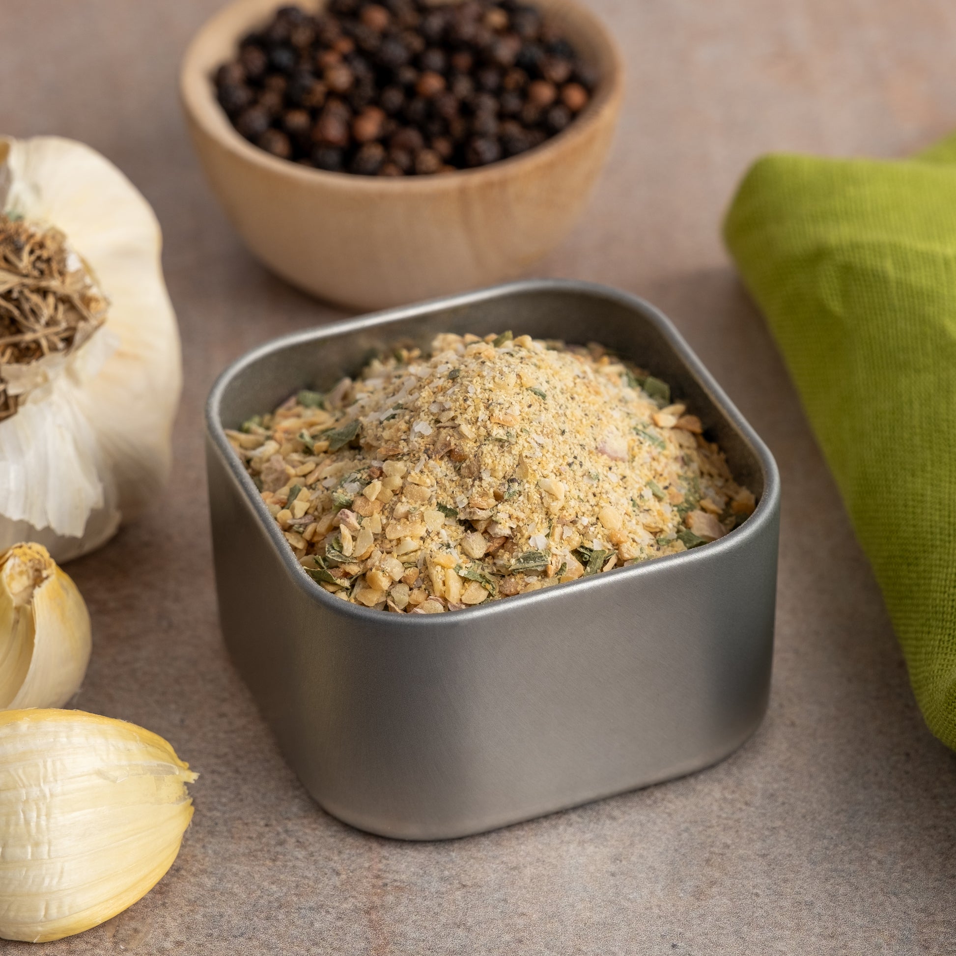 A tin of Onion Obsession Spice Blend rests on a counter near garlic cloves, whole black peppercorns, and a green cloth—ideal for enhancing the flavor of your chicken dishes.