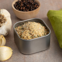 A tin of Onion Obsession Spice Blend rests on a counter near garlic cloves, whole black peppercorns, and a green cloth—ideal for enhancing the flavor of your chicken dishes.