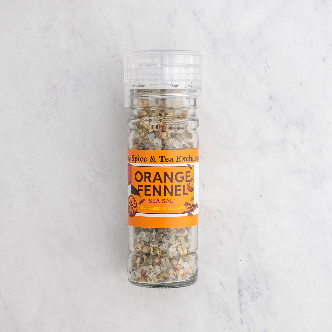 A clear plastic shaker bottle of Spice & Tea Exchange Orange Fennel Sea Salt, showing the salt inside, set against a light gray marble background.