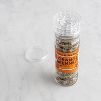 A clear glass spice and tea salt shaker bottle labeled 'Orange Fennel Sea Salt' with its clear lid placed beside it on a light marble surface.