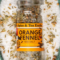 A clear glass jar of Spice & Tea Exchange Orange Fennel Sea Salt, surrounded by coarse white sea salt crystals and dried orange-fennel spices on a dark teal surface.