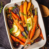 A top-down view of a white rectangular baking dish filled with roasted vegetables, including orange carrots, dark purple beets, sliced lemons, mushrooms, and herbs, with wooden spoons resting on the food.