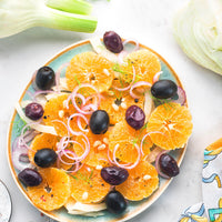 A top-down view of a Mediterranean-style fruit salad in a round ceramic bowl, featuring sliced oranges, black olives, red onion rings, and pine nuts, with green onions and lettuce props on a white marble surface.