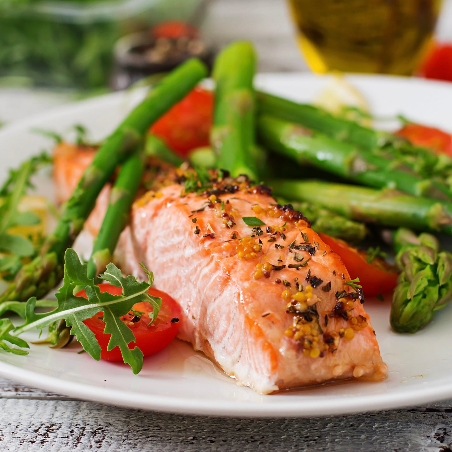A plated salmon fillet with green asparagus, cherry tomatoes, and arugula on a white plate, photographed in a real dining setting with blurred food and table surfaces in the background.