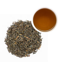 A pile of loose-leaf tea leaves next to a small cup of brewed amber tea on a plain white background.