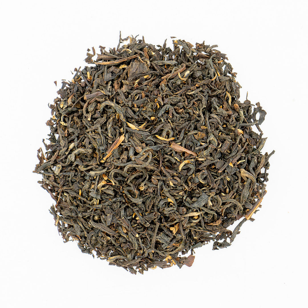 A circular pile of loose-leaf tea leaves, predominantly dark brown and black with lighter tan and yellowish-brown accents, photographed from directly above on a solid white background.